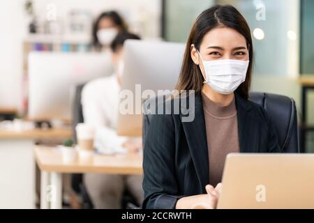 Portrait der asiatischen Büroangestellte Geschäftsfrau tragen Schutzmaske Arbeiten Sie in einem neuen normalen Büro mit interracial Kollegen im Hintergrund Als sozial Stockfoto