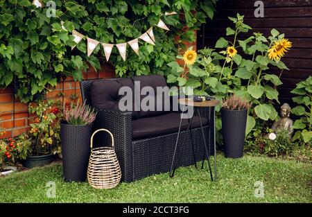 Schwarzer moderner Poly Rattan Gartenmöbelstuhl, Beistelltisch aus Metalldraht. Grüne Weinreben Pflanzen wachsen auf roten Backsteinmauer als dekoratives Element. Holz Stockfoto