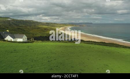 Luftaufnahme der Strandhütte: Grüne Wiese in der Nähe der Meeresbucht und einsame weiße Felsen. Graue Wolken schweben über Ozeanwasser, Sandküste. Ruhige Naturlandschaft in Nordirland. Filmmaterial Stockfoto