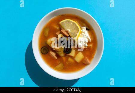 Solyanka-Suppe in einer weißen Schüssel auf blauem Hintergrund. Draufsicht. Berühmte traditionelle ukrainische und russische Salzwürzesuppe mit Zitrone. Stockfoto