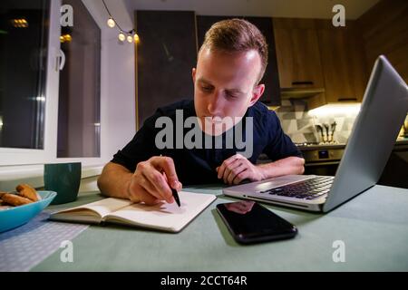 Ein junger Geschäftsmann sitzt an einem Tisch, hält ein Smartphone in der Hand, benutzt einen Laptop und notiert sich in seinem Notebook. Der Typ arbeitet, studiert. Onli Stockfoto