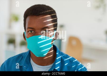 Kopf- und Schulterporträt eines jungen afroamerikanischen Mannes mit Gesichtsmaske und Blick auf die Kamera, die am sonnenbeschienenen Arbeitsplatz im Postpandemic-Büro posiert, Kopierraum Stockfoto