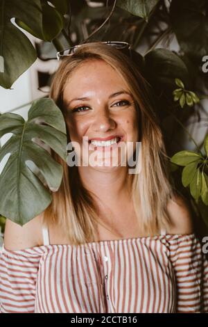 Junge glückliche blonde Frau lächelt durch die Blätter eines monstera Stockfoto