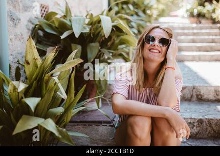 Blonde Frau sitzt lächelnd auf einer Steintreppe mit Pflanzen Stockfoto