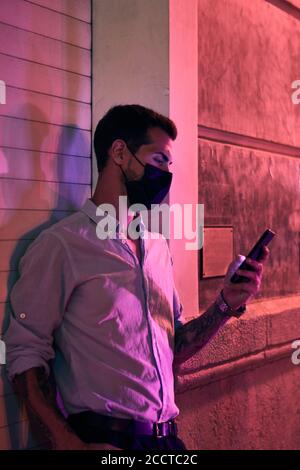 Ein junger Mann mit einer Maske schaut auf sein Handy Nachts Stockfoto