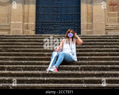 Reife und elegante Frau sitzt auf den Stufen der Kathedrale in Baeza, Jaen, Spanien mit Maske und transparentem Regenschirm Stockfoto