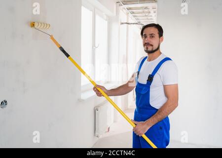 Ein bärtiger Mann in einem blauen Overall malt eine Wand im Flur. Er benutzt eine Walze Stockfoto