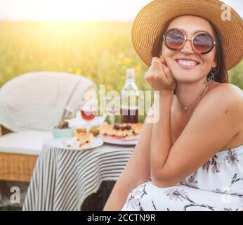 Lächelnd auf Kamera junge aufrichtig Frau in Sonnenbrille und Strohhut gekleidet leichte Sommerkleid sitzen in einem Rattan-Stuhl auf der hohen grünen Wiese Stockfoto