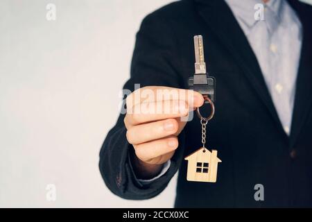Der Typ im Anzug zeigt mir die Schlüssel. Schlüsselanhänger in Form eines Hauses in der Hand eines Mannes. Unerkennbarer Geschäftsmann, der einen Schlüssel anbietet, Nahaufnahme Stockfoto