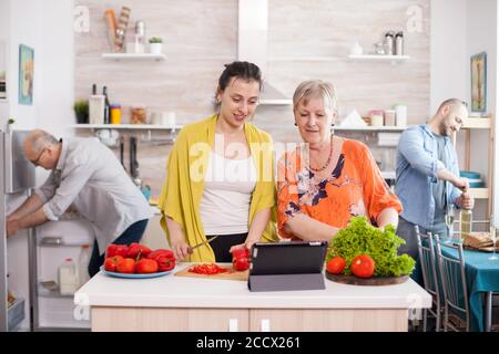 Ältere Frau beobachten Salat Rezept auf dem Tisch beim Kochen zusammen mit ihrer Tochter in der Küche. Mutter Tochter bereitet das Mittagessen vor. Stockfoto