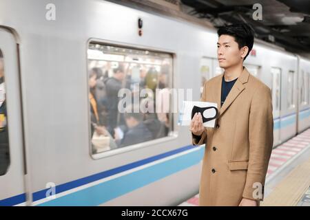 Asiatische Mann hält chirurgische Gesichtsmaske und Karte zu schützen Covid-19 Virus beim Warten auf den nächsten Zug und gehen Zur nächsten Station Stockfoto