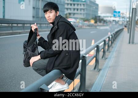 Schöner junger asiatischer Mann tragen Tasche für Reise, während Sitzen auf Metallzaun auf Fußweg Stockfoto