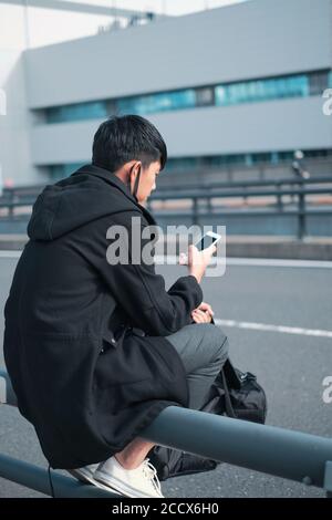 Junge asiatische Mann sitzt auf Straße Barriere wartet auf Bus Und Taxi-Fahrer-Konzept bei der Verwendung von Smartphone Stockfoto