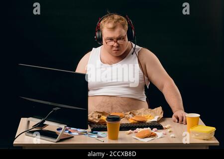 Nachdenklicher nachdenklicher Mann mit Kopfhörern hinter dem Tisch stehend mit Fast Food. Nahaufnahme Porträt. Isolierter schwarzer Hintergrund Stockfoto