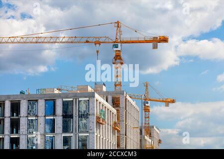 Turmkrane arbeiten auf der Baustelle eines modernen mehrstöckigen Wohngebäudes gegen einen blauen leicht bewölkten Himmel, Kopierraum. Stockfoto