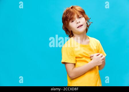 Der rothaarige Junge in einem gelben T-Shirt auf einem blauen Isolierter Hintergrund berührt sich selbst. Stockfoto