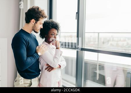 Verärgert afrikanische Frau, die auf das Fenster und ihr kaukasischer Freund tröstet sie, hinter sich stehend und umarmt. Stockfoto