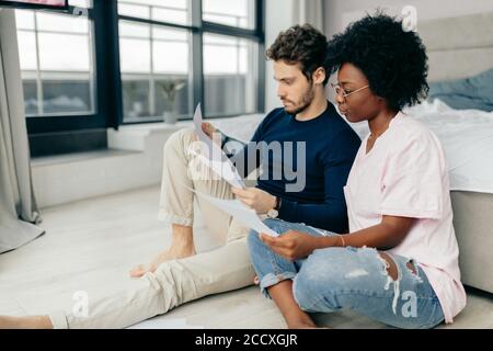 Afrikanische und Kaukasische gemischten Rennen Ehepaar auf dem Boden zu Hause unbezahlte Rechnungen prüfen, Steuern, Schulden, Bank Account Balance sitzen. Es ist ein Stockfoto