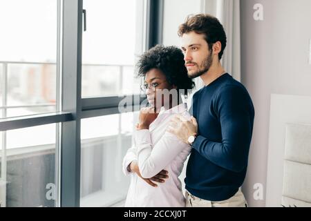 Positive afrikanische Frau umarmt mit ihrem europäischen schönen Brünette Freund, zu Hause entspannen, geben warme Umarmung zueinander, stehen in der Nähe Fenster backgr Stockfoto