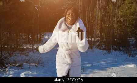 Positive afroamerikanische Dame im weißen Pelzmantel läuft verschneit entlang Winterpark und lächelt gegen hohe Bäume Stockfoto