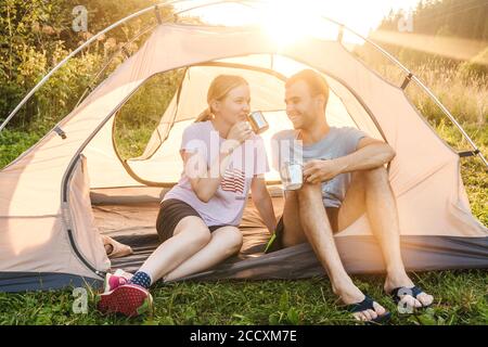 Junge liebende Paar trinken Tee und lächeln im Zelt, Camping in der Natur. Die Sonne geht auf dem Hintergrund auf. Selektiver Fokus. Stockfoto