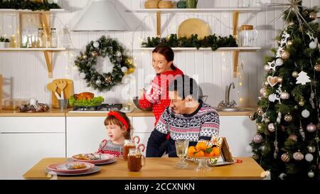 Mutter kocht festliches Abendessen und lächelnd Vater sitzt mit niedlich Mädchen Kind am Tisch in der Küche in der Nähe dekoriert Neujahr Baum Stockfoto