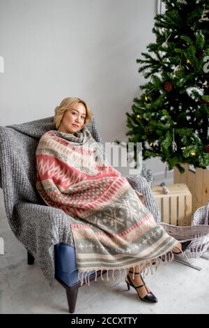Schöne junge blonde Frau in einem gemütlichen weiche Decke gehüllt, sitzt in einem Sessel in der Nähe von Weihnachten Tanne. Stockfoto