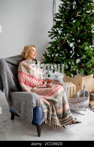 Junge blonde Frau in skandinavischen Ornament karierten sitzen im Stuhl Im Wohnzimmer für Weihnachten dekoriert Stockfoto