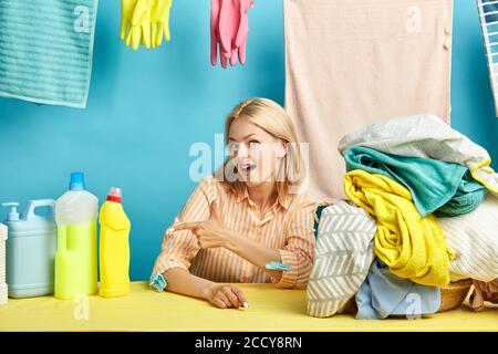 Glückliche junge blonde Frau zeigt auf Spülflüssigkeitsflasche. Blauer Hintergrund, Geste Konzept. Frau gewählt besten effektiven Waschflüssigkeit für Laund Stockfoto