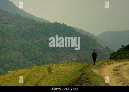 Ein Mann, der auf der Seite der Landstraße steht, mit Blick auf die Reisterrasse, das Tal und die Panchase-Bergkette in der Provinz Gandaki Pradesh in Nepal. Stockfoto