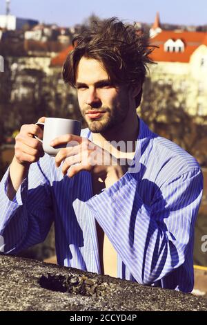 Bärtiger Mann, kurzer Bart. Kaukasische amüsiert Macho mit Schnurrbart und zerzauste Haar in blau gestreiften Schlafanzug holding Tasse oder Becher in Dach auf unscharfen Stadt Hintergrund, morgens Kaffee Konzept Stockfoto