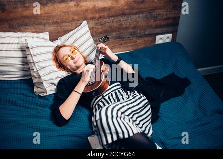 Schöne kaukasische Frau trägt Brillen, mit roten Haaren spielen Ukulele liegen auf dem Bett zu Hause. Schauen Sie sich Kamera Stockfoto