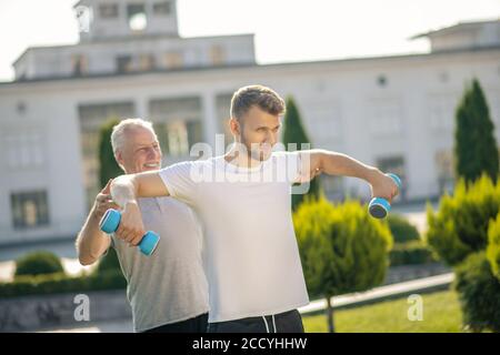 Bärtiger Mann mit Hanteln, grauhaariger Instruktor, der ihm hilft Stockfoto