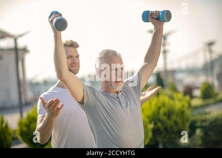 Grauhaariger Mann, der die Hände mit Hanteln hebt, bärtiger Instruktor, der ihm hilft Stockfoto