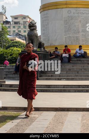 Junger männlicher Mönch, der um Almosen, Essen oder Geld bittet. Gekleidet in traditionelle rote Kleidung, rasierte Kopf. Almschale tragen. Barfuß laufen. Yangon, Myanmar Stockfoto