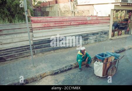 Ägypten, Hurghada. ägyptische Straßenreiniger Senior Mann sitzt auf Bürgersteig trinken Kaffee an alten Zaun Hintergrund Stockfoto