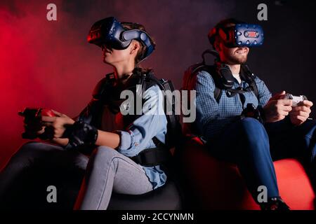 Kaukasische Männer und Frauen sitzen in Headsets, Brillen, Videospiele spielen, Joysticks halten. Rote Neon-Hintergrundbeleuchtung Stockfoto