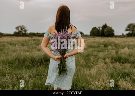 Das Mädchen hält in ihren Händen einen großen Strauß Lila und violette Blüten hinter dem Rücken Stockfoto