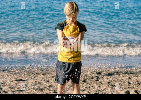 Kleiner Junge am Strand mit Kieseln und Muscheln, die ihm durch die Finger fallen Stockfoto