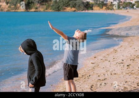 Zwei kleine Jungs, Bruder, stehen am Strand. Der ältere Bruder hat seine Arme in der Luft und blickt mit geschlossenen Augen auf. Stockfoto