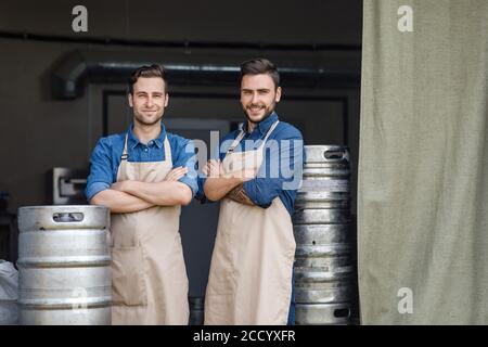 Eigenes kleines Unternehmen. Zwei Jungs kreuzten die Arme über die Brust und stehen in der Nähe von Metallfässern im Brauereilager Stockfoto