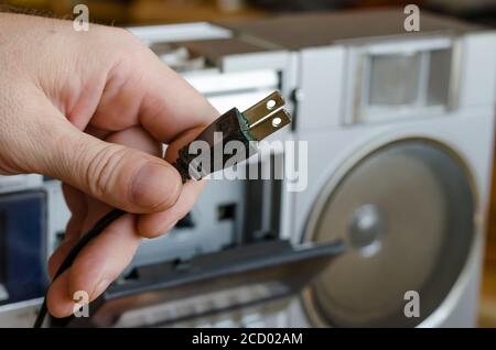 Eine Erwachsene kaukasische männliche Hand hält einen elektrischen Stecker eines Retro-Stereo-Kassettenrecorders. Vintage-Audiogeräte des letzten Jahrhunderts. Selektiver Fokus. Stockfoto