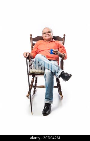 Porträt eines glücklichen reifen indischen oder asiatischen Mann Lesung Ein Buch, während Sie auf einem Schaukelstuhl sitzen und Tee trinken oder Kaffee aus der Tasse Stockfoto