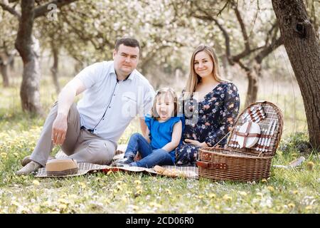 Portrait der glücklichen Familie auf Picknick. Schwangere Mutter, Vater und Tochter sitzen im Frühling blühenden Park. Junge Familie verbringt Zeit zusammen im Urlaub Stockfoto