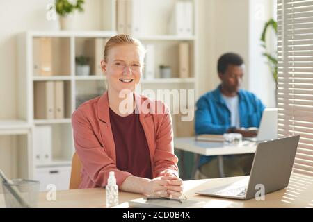 Porträt einer jungen Geschäftsfrau, die mit der Kamera lächelt, während sie am Schreibtisch im Büro mit Händedesinfektionsmittel auf dem Tisch arbeitet, Kopierraum Stockfoto
