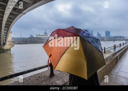 Ein Tourist schaut auf die Stadt London, während sie unter einem bunten Regenschirm unter Blackfriars Bridge vor dem Regen schützen. Stockfoto