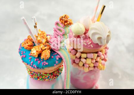 Nahaufnahme von zwei Freak-Shakes, die mit blauen und rosa Donuts, Marshmallow, Bakerüssen, Popcorn und Marmelade auf grauem Hintergrund überziehen; selektiver Fokus Stockfoto