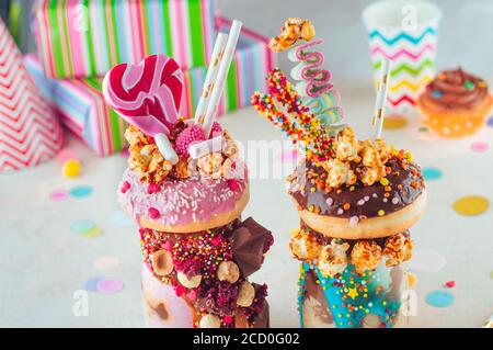 Nahaufnahme von zwei Freak-Shakes mit Donuts, Popcorn und Süßigkeiten auf Party-Hintergrund; selektiver Fokus. Stockfoto