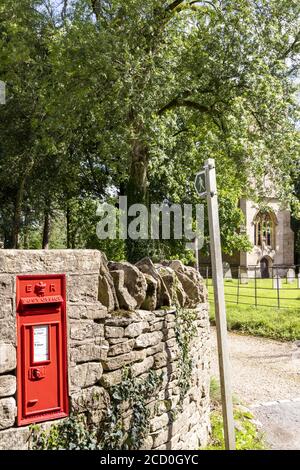 Ein ländlicher Briefkasten und Wegweiser neben der Kirche St. John the Evangelist im Cotswold Dorf Elkstone, Gloucestershire UK Stockfoto