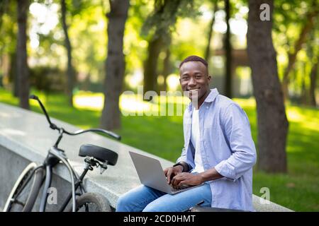 Fröhlicher afroamerikanischer Kerl mit Fahrrad, das am Laptop online im Stadtpark arbeitet, freier Platz Stockfoto